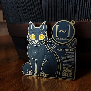 May include: A black and gold metal cat-shaped keychain with the text "Joule Thief Cat" and a QR code. The cat has glowing yellow eyes and a heart on its chest. The keychain is designed to be assembled.