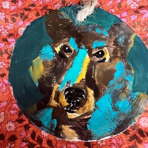 Può includere: Un ornamento rotondo dipinto con il viso di un cane con vernice marrone, gialla e blu. L'ornamento ha una corda per appenderlo.