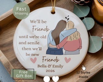 May include: A round white ceramic ornament featuring a gold hanging string. The design shows two illustrated figures with their arms around each other, accompanied by the text We'll be Friends until we're old and senile then we'll be new Friends, Bella & Emily, 2026, and two small pink hearts.