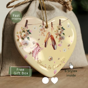 May include: A heart-shaped ceramic ornament with a pale yellow background featuring a ballet dancer in a pink dress, ballet slippers hanging from a string, and floral accents. The text reads Olivia As Clara 2026 in a cursive script font.