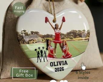 May include: A heart-shaped ceramic ornament featuring a printed photograph of three cheerleaders in red and white uniforms on a green field. The ornament includes the name OLIVIA and the year 2026 in black text, with a gold hanging cord.