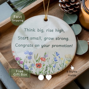 May include: A round white ceramic ornament featuring a gold hanging string and a colorful floral print at the bottom. The text reads: Think big, rise high, Start small, grow strong. Congrats on your promotion! Two small white charms hang below.