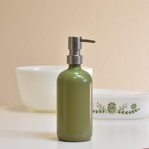 May include: A green glass soap dispenser with a silver metal pump. The dispenser is cylindrical and has a glossy finish. The background includes white bowls and a light-colored wall.