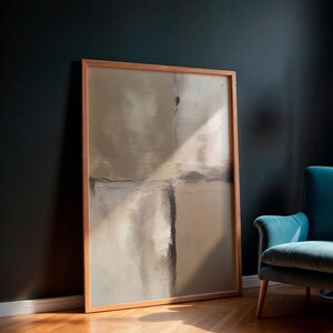 May include: Large framed abstract art print with neutral tones of beige, gray, and brown. The artwork is in a light wood frame and leans against a dark wall. A teal armchair sits nearby on a hardwood floor.