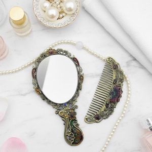 May include: An antique-style hand mirror and comb set with a decorative floral design in gold, red, and green. The oval mirror and comb are on a white marble surface, with a pearl necklace and perfume bottles.