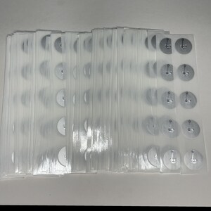 May include: A stack of clear plastic sheets, each containing several small, silver-colored metal discs with attached pins. The discs are arranged in rows and columns, and the sheets are stacked neatly together.