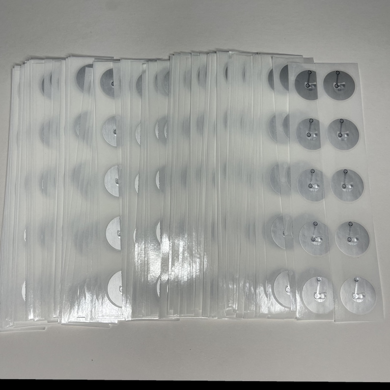 May include: A stack of clear plastic sheets, each containing several small, silver-colored metal discs with attached pins. The discs are arranged in rows and columns, and the sheets are stacked neatly together.