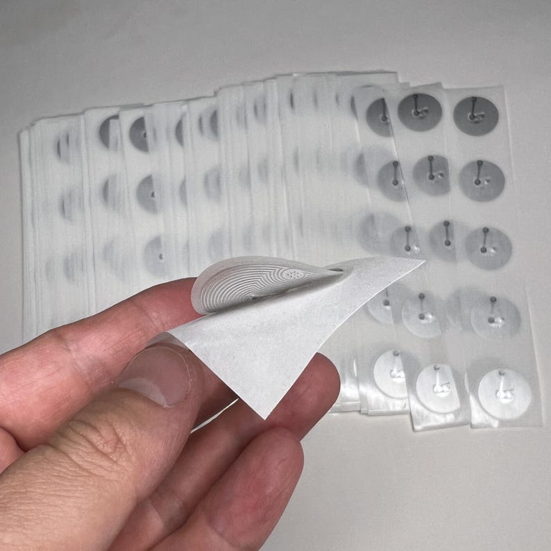 May include: A stack of clear, adhesive RFID labels with a peel-off corner. The labels are round with a small antenna design and are on a clear backing. The corner of one label is peeled back, revealing the antenna.