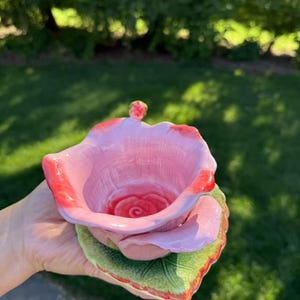 Puede incluir: Un cuenco de cerámica con forma de rosa, en tonos rosa y rojo. El cuenco se asienta sobre una base verde con forma de hoja. El interior del cuenco presenta un centro de rosa detallado. El cuenco se sostiene en una mano.