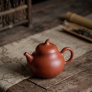 May include: A round, reddish-brown clay teapot featuring a curved spout, a circular handle, and a lid topped with a small, pointed finial. The surface of the teapot has a textured, matte finish, and the overall design is simple and functional.