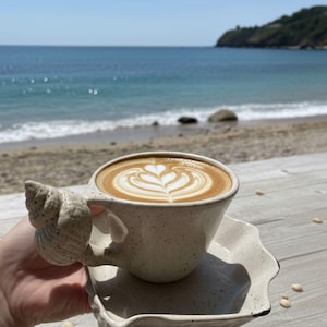 May include: A ceramic coffee cup and saucer set with a seashell handle, filled with latte art. The cup and saucer are speckled beige. The latte art features a heart design. The background shows a beach and ocean.