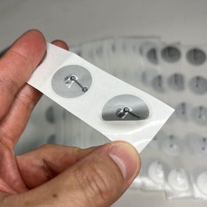 May include: A strip of white adhesive labels with two circular RFID tags. Each tag features a gray spiral antenna and a small black square. The background shows additional labels, suggesting a bulk product.