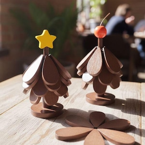 Könnte beinhalten: Weihnachtsbaumschmuck aus Holz. Zwei Bäume mit braunen Holzblättern und einem gelben Stern oder einer roten Kirsche oben. Eine Holzblume ist ebenfalls vorhanden. Die Artikel befinden sich auf einer hellen Holzoberfläche.