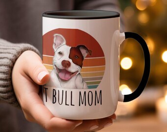 May include: White ceramic mug with a black handle. The mug features a colorful retro sunset design with a white and brown pit bull dog in the center. The text "Pit Bull Mom" is printed in black below the dog.