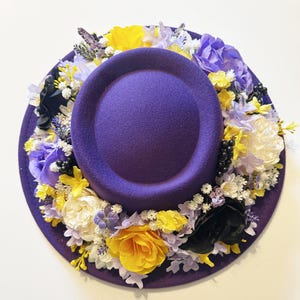 May include: A purple felt hat with a wide brim decorated with a wreath of yellow, white, and purple flowers. The flowers are arranged in a circle around the hat's crown.