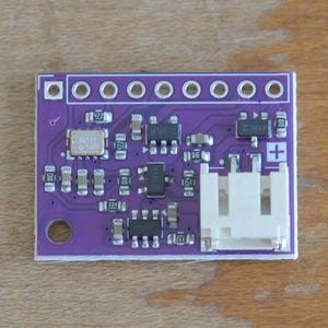 May include: A small, rectangular purple circuit board with various electronic components, including chips, resistors, and a white connector. The board has a series of holes along the top edge and a single hole on the bottom left corner. The components are arranged in an organized manner.