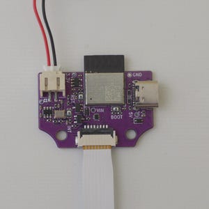 May include: A purple printed circuit board featuring a central metallic module, a USB-C port on the right, and a white connector with red and black wires on the left. A flat white ribbon cable is attached to the bottom, with labels including 3.3V, GND, VIN, BOOT, and INT printed on the board surface.