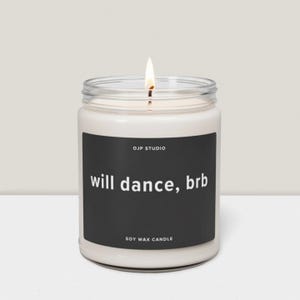 Könnte beinhalten: Eine weiße Sojawachskerze in einem transparenten Glas mit einer brennenden Docht. Das schwarze Etikett trägt den weißen Schriftzug "will dance, brb", darüber "OJP STUDIO" und darunter "SOY WAX CANDLE".