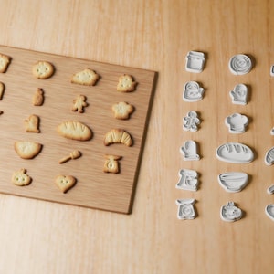 Könnte beinhalten: Eine Sammlung weißer Kunststoff-Ausstechformen in verschiedenen küchenbezogenen Motiven wie Mixer, Nudelholz und Brotlaib, präsentiert neben den passenden gebackenen Keksen auf einem rechteckigen Holzbrett.
