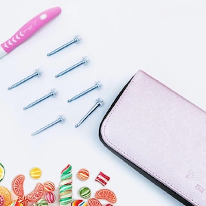 May include: A pink crochet hook and several silver screws are arranged on a white surface. A pink case with a black border is on the right. Colorful candies are scattered at the bottom.
