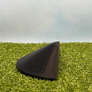 May include: A black, triangular object with a ridged surface, resting on a bed of green artificial turf. The object has a curved shape and appears to be made of a solid material. The background is a gradient of light blue and white.