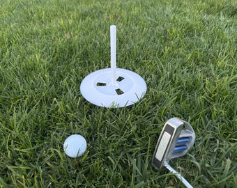 May include: A white plastic golf ball holder with a white golf ball and a silver putter on green grass.