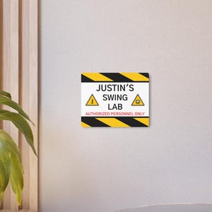 May include: A rectangular sign with a black and yellow striped border. The sign reads "Justin's Swing Lab" with warning symbols and the text "Authorized Personnel Only."