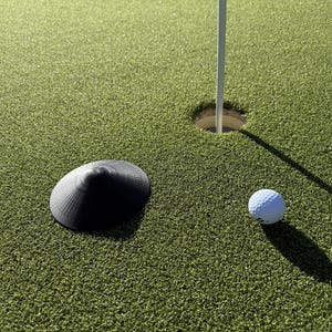 May include: A golf course green with a white golf ball, a black ball marker, a golf hole, and a white flagstick. The green is covered in short, artificial turf. The ball is near the hole.