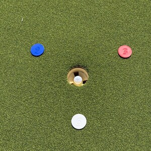 May include: A white golf ball sits inside a hole on a green putting surface. Three circular markers are arranged around the hole: a blue marker with the number 2, a red marker with the number 3, and a white marker with the number 6.