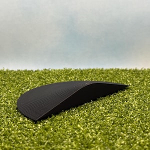 May include: A black, curved object with a ridged surface, resting on a bed of green artificial grass. The object has a smooth, rounded shape and appears to be made of a solid material. The background is a gradient of blue and white.