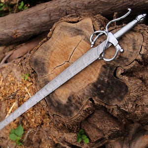 Peut inclure: Une épée argentée avec une lame à motifs et une garde ornée repose sur une souche d'arbre vieillie. La poignée de l'épée présente un design complexe et la lame a un aspect texturé. L'arrière-plan comprend du bois et du feuillage vert.