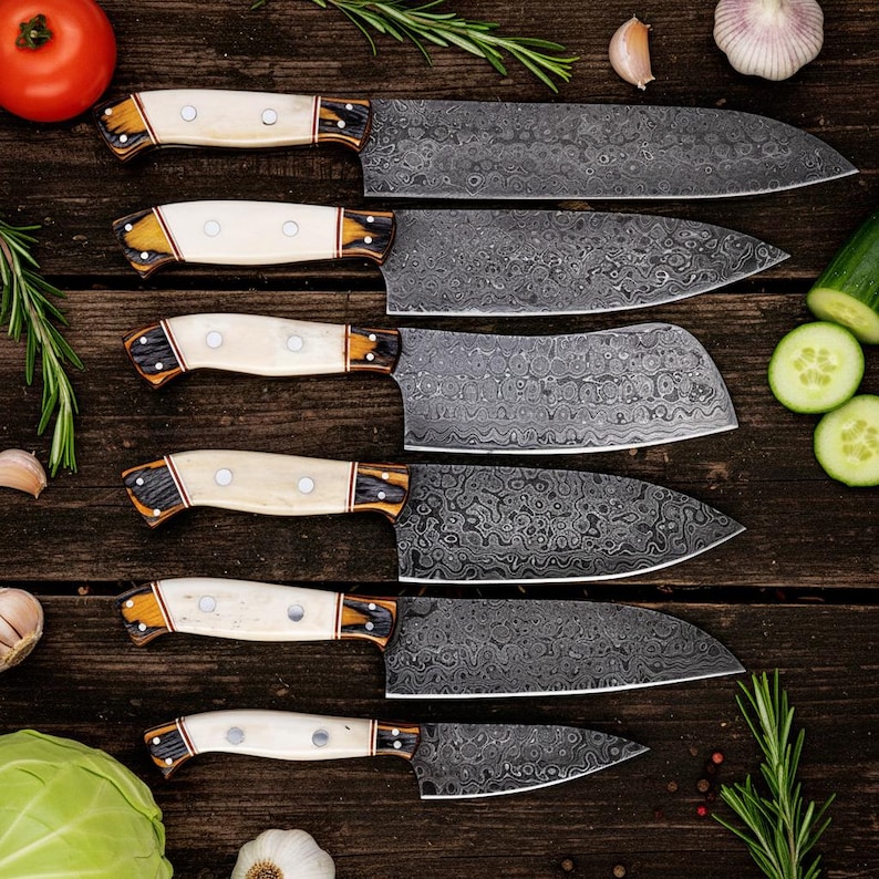 May include: An overhead shot showcases a set of six Damascus steel kitchen knives with cream-colored handles and intricate blade patterns. The knives are arranged on a wooden surface, surrounded by fresh vegetables and herbs, suggesting a culinary theme.