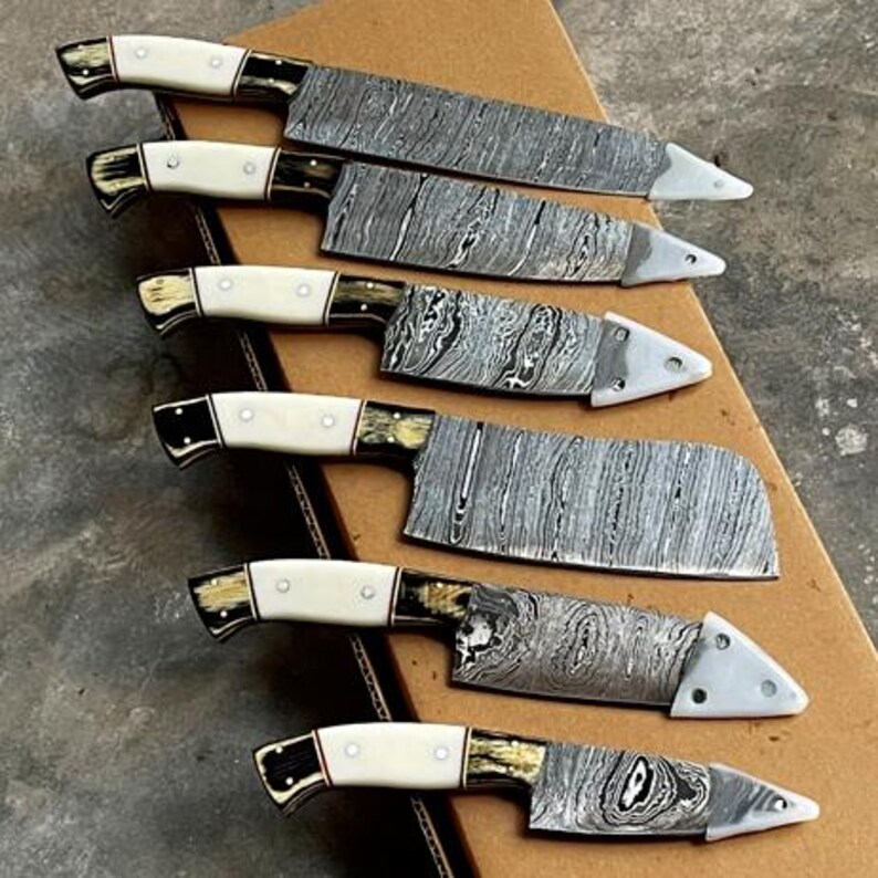 May include: A collection of seven kitchen knives with patterned steel blades and cream-colored handles. The handles have black and gold accents. The blades have a unique, swirling pattern. The knives are arranged on a cardboard surface.