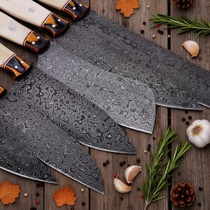 May include: A set of five Damascus steel kitchen knives with patterned blades and cream-colored handles. The blades have a unique, swirling design. The handles are accented with dark brown and gold details. The knives are arranged on a wooden surface.