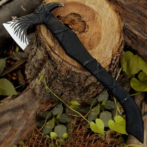 Könnte beinhalten: Eine schwarze Axt mit einem lederumwickelten Stiel. Der Axtkopf hat ein dekoratives Muster und liegt auf einem Baumstumpf.