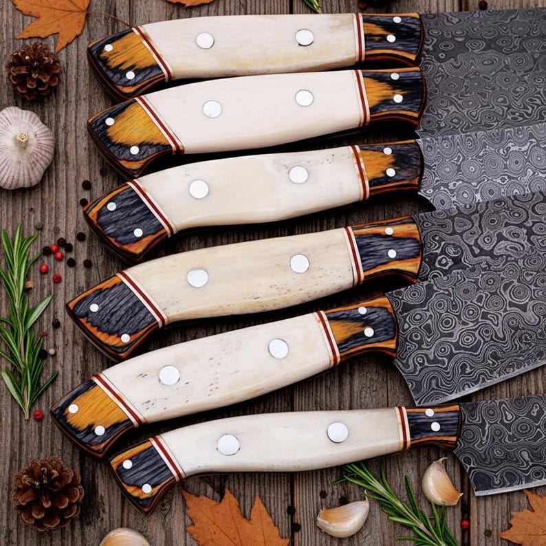 May include: Six Damascus steel knives with cream-colored handles and decorative wood accents. The handles are secured with silver rivets. The blades have a unique patterned design. The knives are arranged on a wooden surface with herbs and spices.