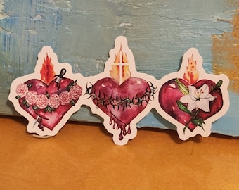 May include: Three watercolor stickers depicting the Sacred Hearts of Jesus and Mary. Each heart is pierced with a symbol of their suffering: a sword, a crown of thorns, and a lily. The hearts are surrounded by flames and a halo.
