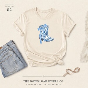 May include: A cream-colored short-sleeve t-shirt featuring a central graphic of two cowboy boots decorated with a blue and white floral pattern and a blue ribbon bow. The text THE ART FILE 02 and THE DOWNLOAD DWELL CO. ARTWORK PREVIEW ON APPAREL appears.