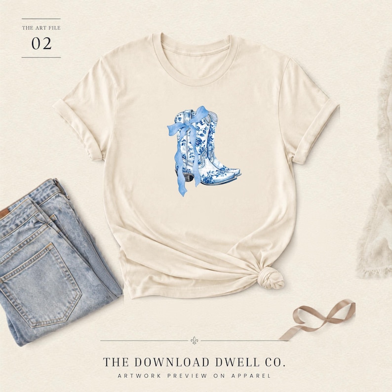 May include: A cream-colored short-sleeve t-shirt featuring a central graphic of two cowboy boots decorated with a blue and white floral pattern and a blue ribbon bow. The text THE ART FILE 02 and THE DOWNLOAD DWELL CO. ARTWORK PREVIEW ON APPAREL appears.