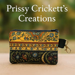 May include: A small, rectangular coin purse with a black zipper and a black wrist strap. The purse features a colorful floral pattern in yellow, black, orange, and blue. The text "Prissy Crickett's Creations" is at the top.