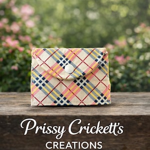 May include: A small, plaid fabric wallet with a cream background and a pattern of red, yellow, and blue lines. The wallet has a flap secured by a white button. The text "Prissy Crickett's CREATIONS" is below the wallet.