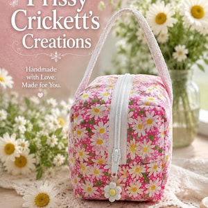 May include: A quilted fabric pouch featuring a pink background with a white daisy pattern, a white zipper closure, and a white handle. A white daisy-shaped charm is attached to the zipper pull. The text reads Prissy Crickett's Creations, Handmade with Love. Made for You. Unique Designs, Quality Craftsmanship, Made to Brighten Your Day.