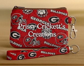 May include: A red zippered pouch and matching wrist strap featuring a repeating pattern of football helmets, the letter "G", and the word "Bulldogs". The pouch and strap have silver-colored hardware and the text "Prissy Crickett's Creations".