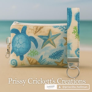 May include: A small, zippered pouch and matching wrist strap featuring a sea-themed pattern. The design includes blue sea turtles, starfish, and seashells on a cream-colored background. The wrist strap has a silver-colored metal ring.