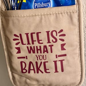 May include: A beige oven mitt with the text "Life is what you bake it" in maroon lettering. The oven mitt has a pocket on the front with a blue Pillsbury package inside.