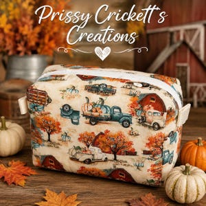 May include: A rectangular fabric pouch featuring a cream background with a repeating autumn pattern of red barns, teal and white pickup trucks, orange pumpkins, and fall foliage. The pouch has a white zipper closure and side fabric tabs.