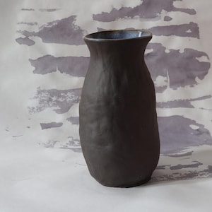 May include: A dark brown ceramic vase with a textured surface. The vase has a slightly flared rim with a blue interior. The background features a white surface with gray watercolor-like patterns.