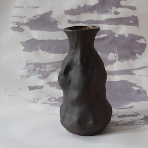 May include: A dark brown ceramic vase with a textured surface. The vase has a narrow neck and a rounded body. The background is a light gray and white abstract pattern. The vase is approximately 8 inches tall.