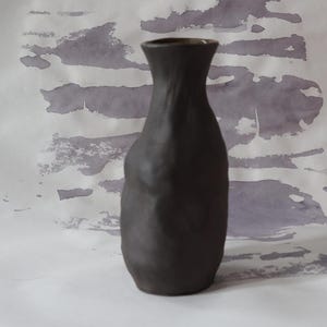 May include: A dark gray ceramic vase with a narrow neck and a slightly textured surface. The vase has a rounded body and stands on a white surface with gray watercolor-like patterns.