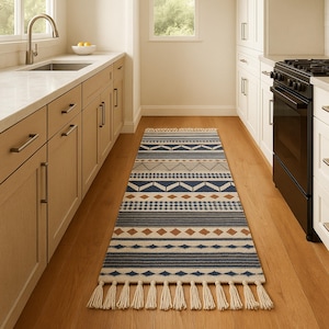 May include: A rectangular kitchen runner rug with a geometric pattern in navy blue, gray, and tan. The rug features diamond, triangle, and stripe designs. It has a cream-colored fringe on both ends. The rug is placed on a hardwood floor.